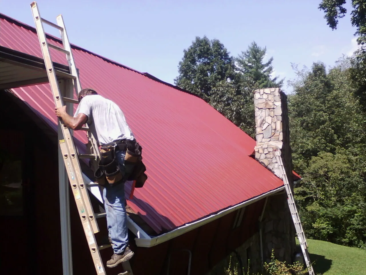 Roofer on a ladder beside a red metal roof and stone chimney — Soffit & Fascia Repair by Roofing Brothers in Victor